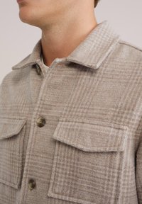 Heled pruun villane särk-jakk, millel on ruuduline muster, kahe rinnatasku ja nööbikinnitustega. Pehme tekstuur ja klassikaline krae.