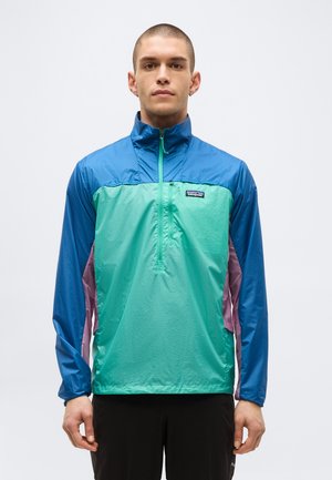 Férfi, kék és türkizszínű, félcipzáras Patagonia dzsekit visel lila oldalpanelekkel és fekete nadrággal, előre néz egy fehér háttér előtt.