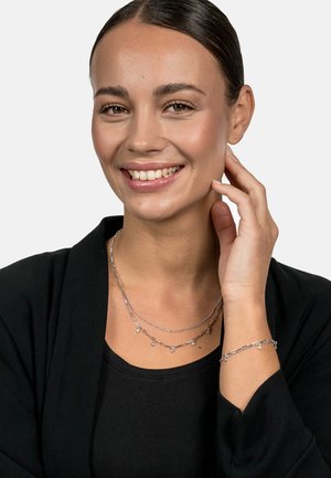 Silver layered necklace with flat links and small charms, paired with a matching bracelet, worn over a black top. Smooth texture, reflective finish.
