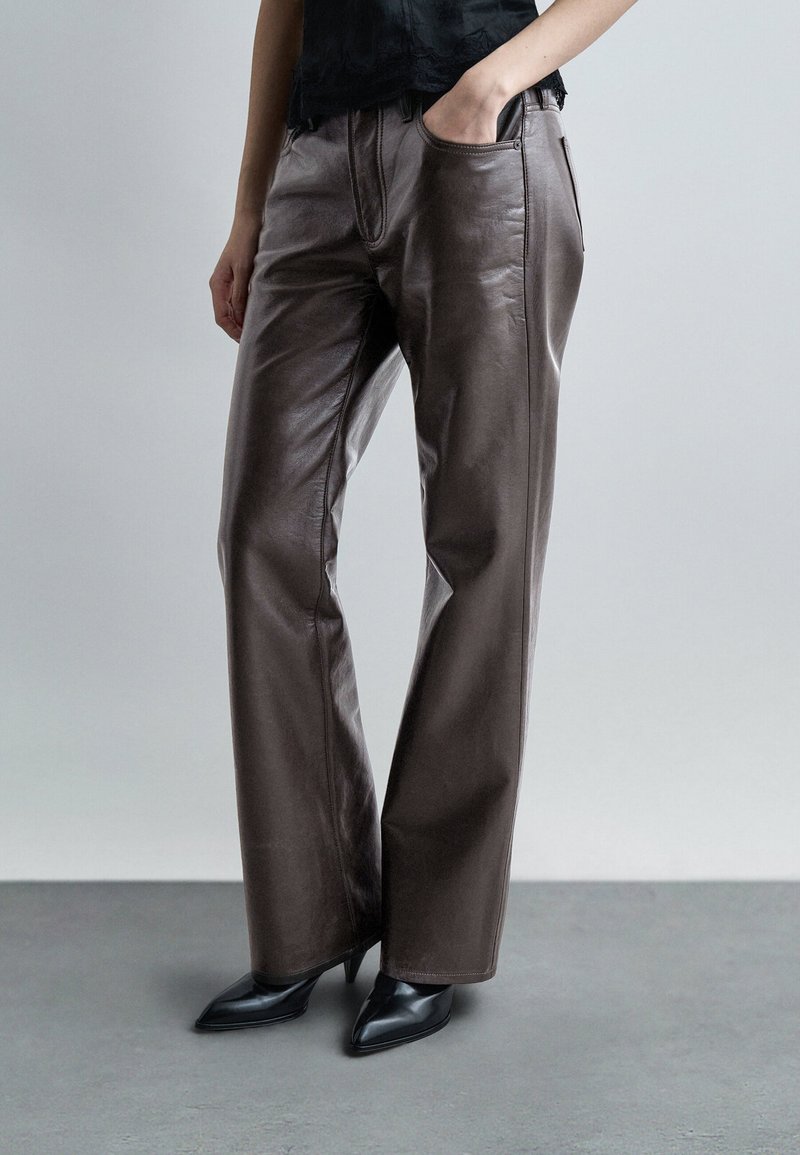 Oseba, ki nosi temno rjave usnjene hlače in črne čevlje z visokimi petami ter zašiljenimi konicami, stoji z eno roko v žepu na nevtralni podlagi.