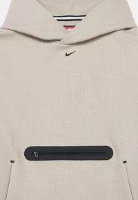 Μπεζ φούτερ με κουκούλα, με μαύρο λογότυπο Nike, μπροστινή τσέπη με φερμουάρ και μαλακή, λεία υφή. Διαθέτει κουκούλα με κορδόνι και ριγέ εσωτερική επένδυση.
