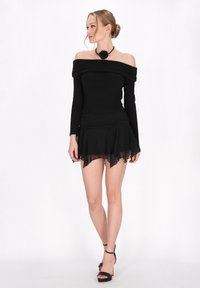 Zwart off-shoulder top met lange mouwen, gecombineerd met een gelaagde zwarte chiffon rok. Opvallende rozenhanger ketting en zwarte hakken.