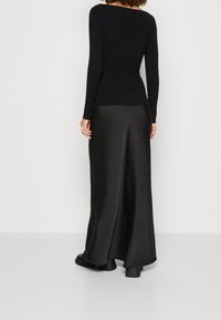 Frau in einem schwarzen, gerippten Langarmshirt, langer schwarzer Satin-Rock und schwarzen Stiefeln, die mit dem Rücken zur Kamera steht.