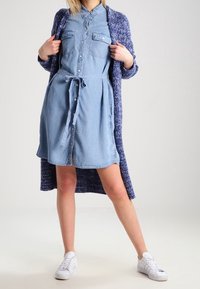 Ljusblå denimskjolklänning med knyt i midjan, ihop med en mörkblå texturerad kofta. Vita sneakers fulländar outfiten.