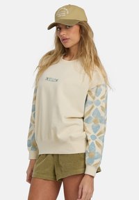 Sudadera beige con mangas de un patrón azul y dorado, puños acanalados y un pequeño logotipo bordado. Combinada con pantalones cortos de pana en color oliva.