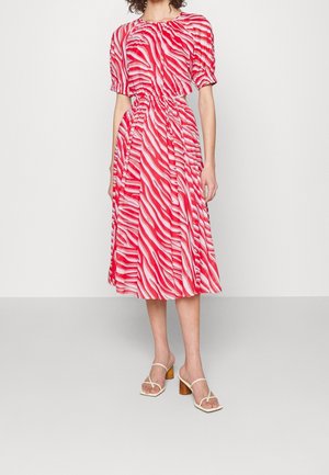 Vrouw die een midi-jurk draagt met rood-witte zebraprint en opgeblazen mouwen, en beige sandalen met hakken, staand tegen een effen achtergrond.