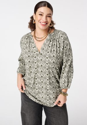 Femme souriante aux cheveux bouclés portant un chemisier vert à motifs, un pantalon noir, des boucles d'oreilles en anneaux dorés, des colliers superposés et des bracelets sur un fond blanc.