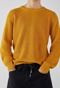 Gelber Strickpullover mit Rundhalsausschnitt, gerippter Textur und langen Ärmeln, kombiniert mit schwarzen Jeans mit einem Kordelzugbund.