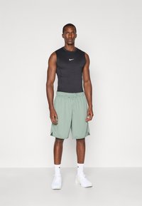 Schwarzes ärmelloses Sportoberteil mit Nike-Logo, kombiniert mit hellgrünen Sportshorts. Das Modell trägt weiße Schuhe mit passenden Socken.