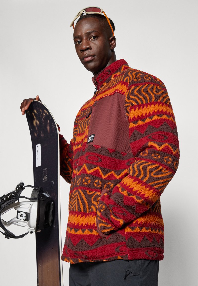 Red and orange patterned fleece jacket with a brown panel, side pocket, and high collar; model holding a snowboard with bindings.