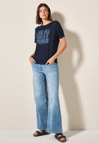 T-shirt à manches courtes bleu marine avec un design graphique abstrait bleu sur le devant, associé à un jean large bleu clair et des sandales sombres.
