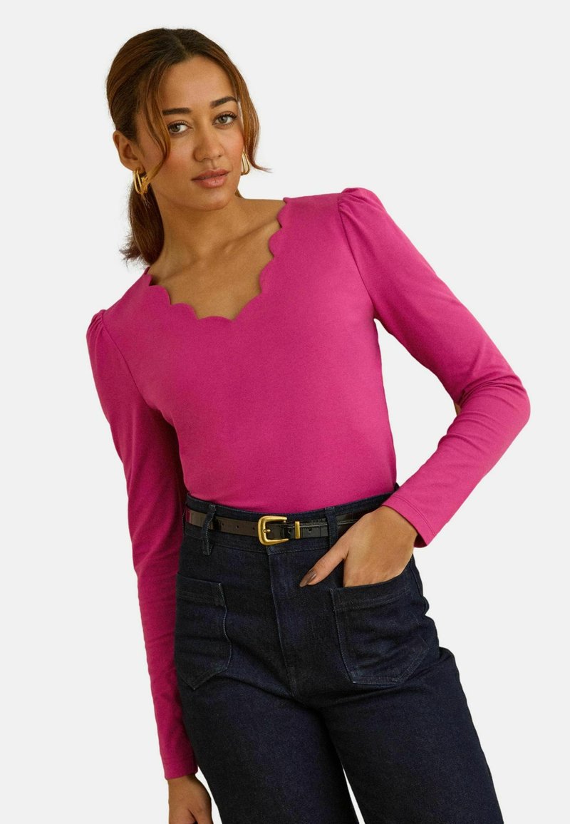 Felroze top met lange mouwen, een geschulpte halslijn en geplooide schouders, gecombineerd met donker denim broek en een zwarte riem.