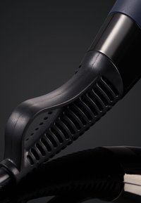 Gros plan sur la poignée d'un sèche-cheveux noir avec grip texturé et design ventilé sur une surface réfléchissante sombre.