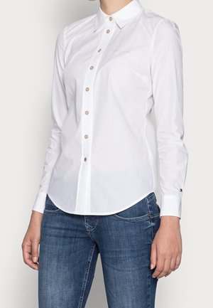 Person iført en tætsiddende hvid skjorte med lange ærmer og blå jeans, stående mod en ensfarvet baggrund.