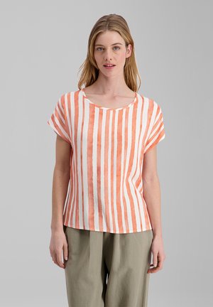 Frau trägt ein weißes Kurzarm-Top mit vertikalen orangefarbenen Streifen und lockere hellbraune Hose, steht vor einem schlichten grauen Hintergrund.