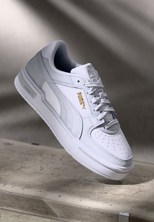 White sneaker with smooth leather material, perforated toe, and a textured sole. Features subtle gold branding on the side.