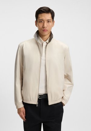 Helles beiges Jackett mit einem Reißverschluss vorne, elastischen Bündchen und einem gemusterten Kragen. Getragen über einem weißen Poloshirt, kombiniert mit dunklen Hosen.