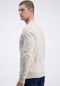 Beżowa bluza z okrągłym dekoltem, z prążkowanymi mankietami i pasem, ozdobiona metką na lewym rękawie. Gładka tkanina o dopasowanym kroju.