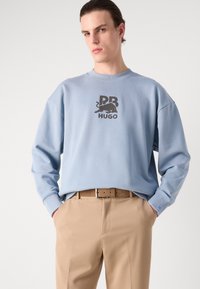 Sudadera azul claro de corte relajado, con gráficos impresos en negro y texto en la parte frontal. Combinada con pantalones color caqui y un cinturón marrón.