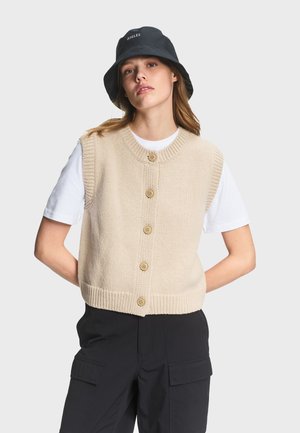 Donna che indossa un gilet lavorato a maglia beige abbottonato sopra una t-shirt bianca, cappello a secchiello nero e pantaloni cargo neri, in piedi con le mani dietro la schiena.