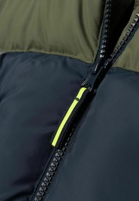 Veste zippée avec un haut vert olive et un bas bleu marine. Comprend une fermeture éclair noire avec un tirant jaune lumineux et un tissu texturé.