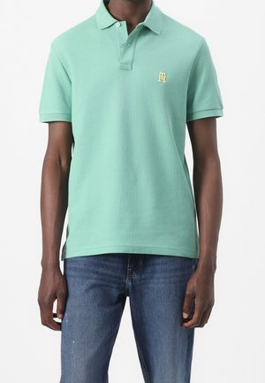 Grøn polo-shirt lavet af tekstureret stof med en krave, tre knapper og et lille gult logo på venstre bryst. Bæres med blå jeans.