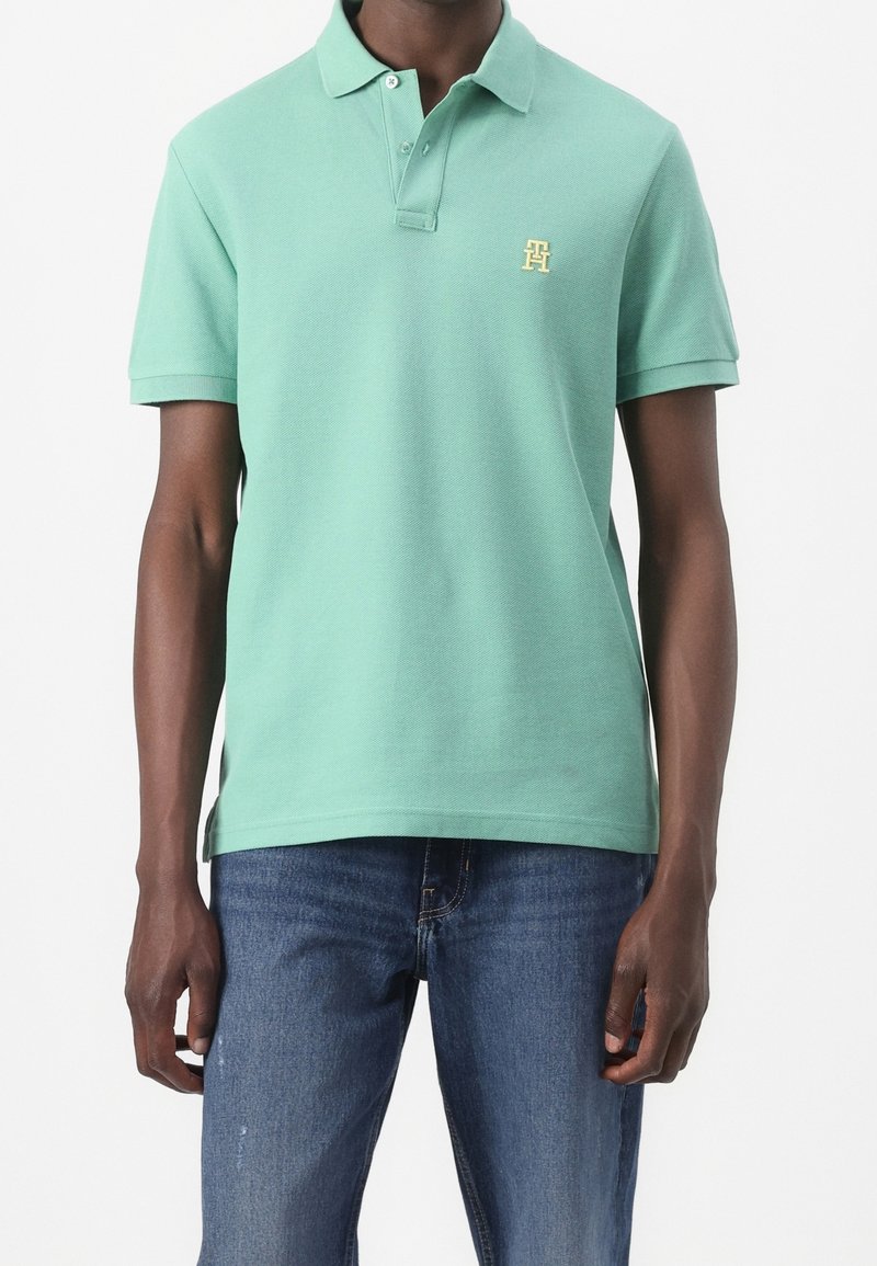 Polo vert en tissu texturé avec un col, trois boutons et un petit logo jaune sur la poitrine gauche. Porté avec un jean bleu.