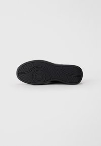 Sole di sneaker nera con superficie in gomma testurizzata, caratterizzata da un logo circolare e un motivo a zigzag. Forma piatta e arrotondata, senza accenti visibili.