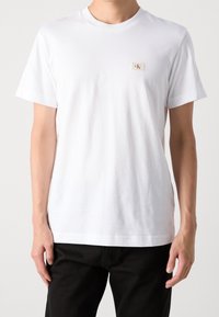 Uomo che indossa una maglietta bianca a maniche corte con un piccolo logo beige di Calvin Klein sul petto, abbinata a pantaloni neri, su uno sfondo neutro.