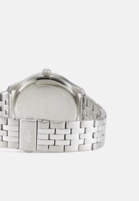 Ceas de mână argintiu cu o brățară metalică cu finisaj texturat și un carcasă rotundă. Include o coroană vizibilă și un logo gravat pe clapetă.
