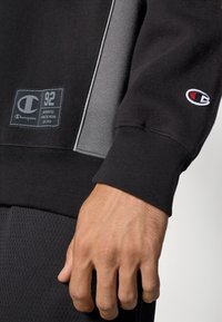 Sudadera negra con panel gris, puños de canalé y un parche con logo. Presenta una tela texturizada y un brillo sutil, enfatizando el diseño atlético.
