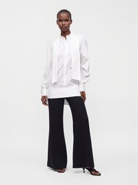 Personne debout portant un chemisier blanc à manches longues avec des détails à nouer et un pantalon noir à jambes larges avec des talons ouverts noirs.