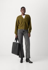 Ελιές πράσινο cardigan με V-λαιμόκοψη και κλείσιμο με κουμπιά, συνδυασμένο με γκρι φαρδιά παντελόνια και μαύρες μπότες στον αστράγαλο, κρατώντας μια δομημένη μαύρη τσάντα.