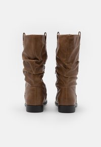 Bottes montantes en cuir marron avec texture froncée, bout arrondi et petit talon noir ; équipées de deux languettes pour un enfilage facile.