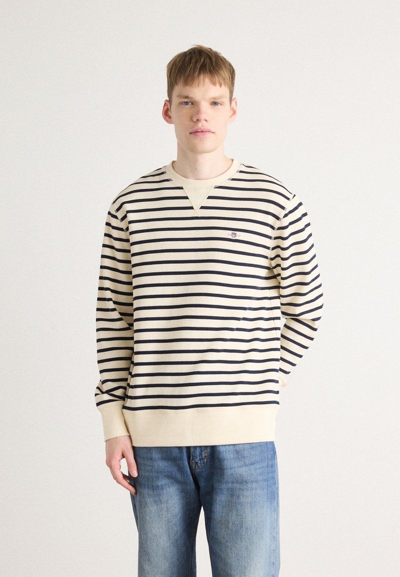 Kremfarget og marineblå stripete genser med ribbet tekstur, v-halsdesign og liten brodert logo på brystet. Brukt med blå jeans.