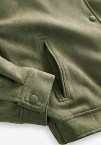 Olivově zelená bunda vyrobená z měkkého materiálu má manžety na knoflík a boční kapsy. Materiál má hladký, semišový povrch.