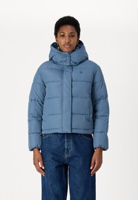 Giacca imbottita azzurro chiaro con colletto alto e cappuccio staccabile, dotata di chiusura con zip e bottoni, abbinata a jeans blu a gamba larga.