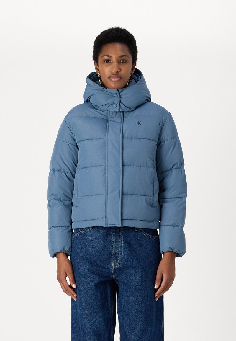 Giacca imbottita azzurro chiaro con colletto alto e cappuccio staccabile, dotata di chiusura con zip e bottoni, abbinata a jeans blu a gamba larga.