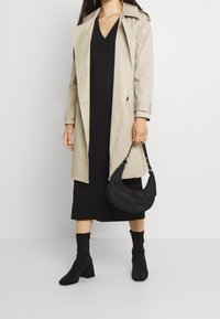 Cappotto trench beige sopra un abito midi nero a coste, abbinato a stivaletti neri con tacco e una borsa a forma di mezza luna nera.