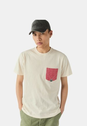 T-shirt di cotone color crema con una tasca rossa, caratterizzata da un motivo a diamante. Il modello indossa un cappellino grigio scuro con logo ricamato.