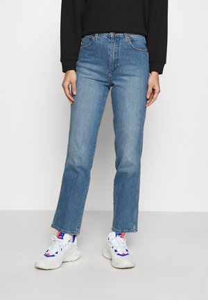 Personne portant un jean bleu à jambes droites, des baskets blanches avec des détails bleus et rouges, et un haut noir à manches longues, debout sur un sol gris.