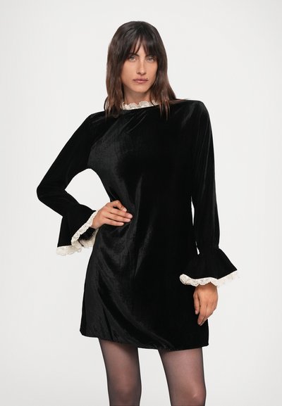Black velvet dress with a high neckline and flared sleeves, trimmed with white lace at the cuffs. Smooth texture, above-the-knee length.