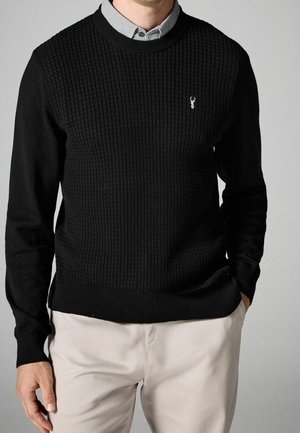 Homme portant un pull noir texturé avec un petit logo de cerf blanc sur la poitrine, par-dessus une chemise à col à motifs, et un pantalon beige clair.