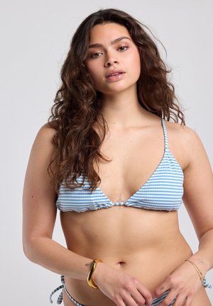 Mujer con cabello largo y rizado, vistiendo un bikini a rayas azul y blanco, sosteniendo los lazos de la parte inferior del bikini, con una pulsera dorada en la muñeca.