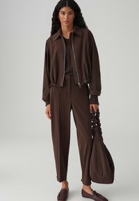 Bruine bomberjack met rits over een donkerbruine trui en bijpassende broek. De outfit wordt aangevuld met een slouchy handtas en bruine loafers.