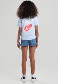 T-shirt di cotone azzurro chiaro con una grafica di astice rosso e testo sul retro, abbinato a pantaloni corti di denim e sneakers bianche con accenti neri.