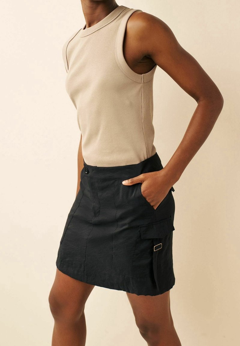 Top sans manches beige avec texture côtelée, associé à une jupe cargo noire dotée de poches et d'un détail de boucle sur le côté.