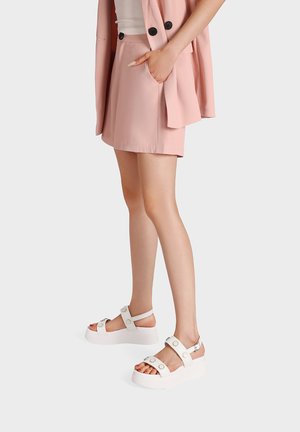 Donna che indossa pantaloncini rosa chiaro su misura e blazer abbinato con sandali bianchi con plateau, in piedi davanti a uno sfondo semplice.