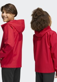 Deux jeunes portant des vestes à capuche rouges sont dos à dos devant un fond uni.