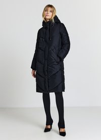 Cappotto lungo nero in stile piumino con cappuccio, chiusura a bottoni e motivo di cuciture diagonali; abbinato a collant neri e tacchi. Texture liscia.
